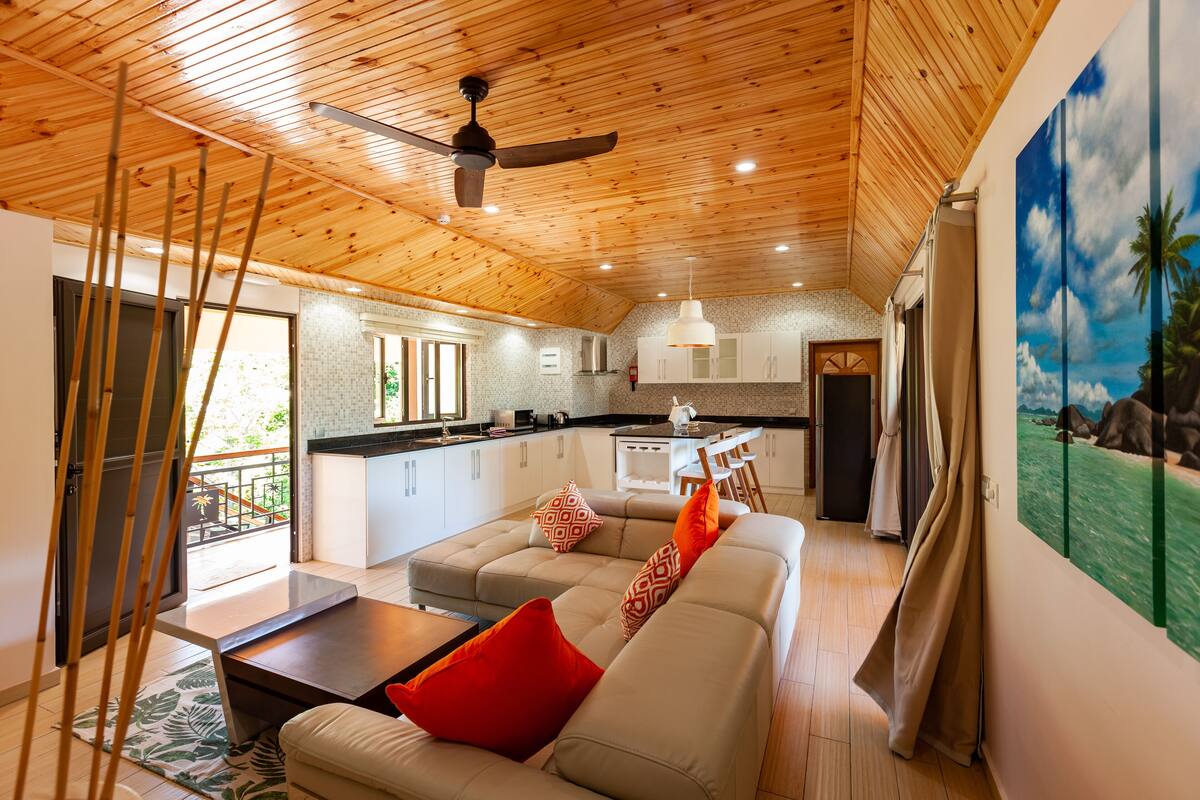 A modern kitchen with white cabinets, a dining area with a white table and chairs, and a living room with a beige sofa and red cushions.