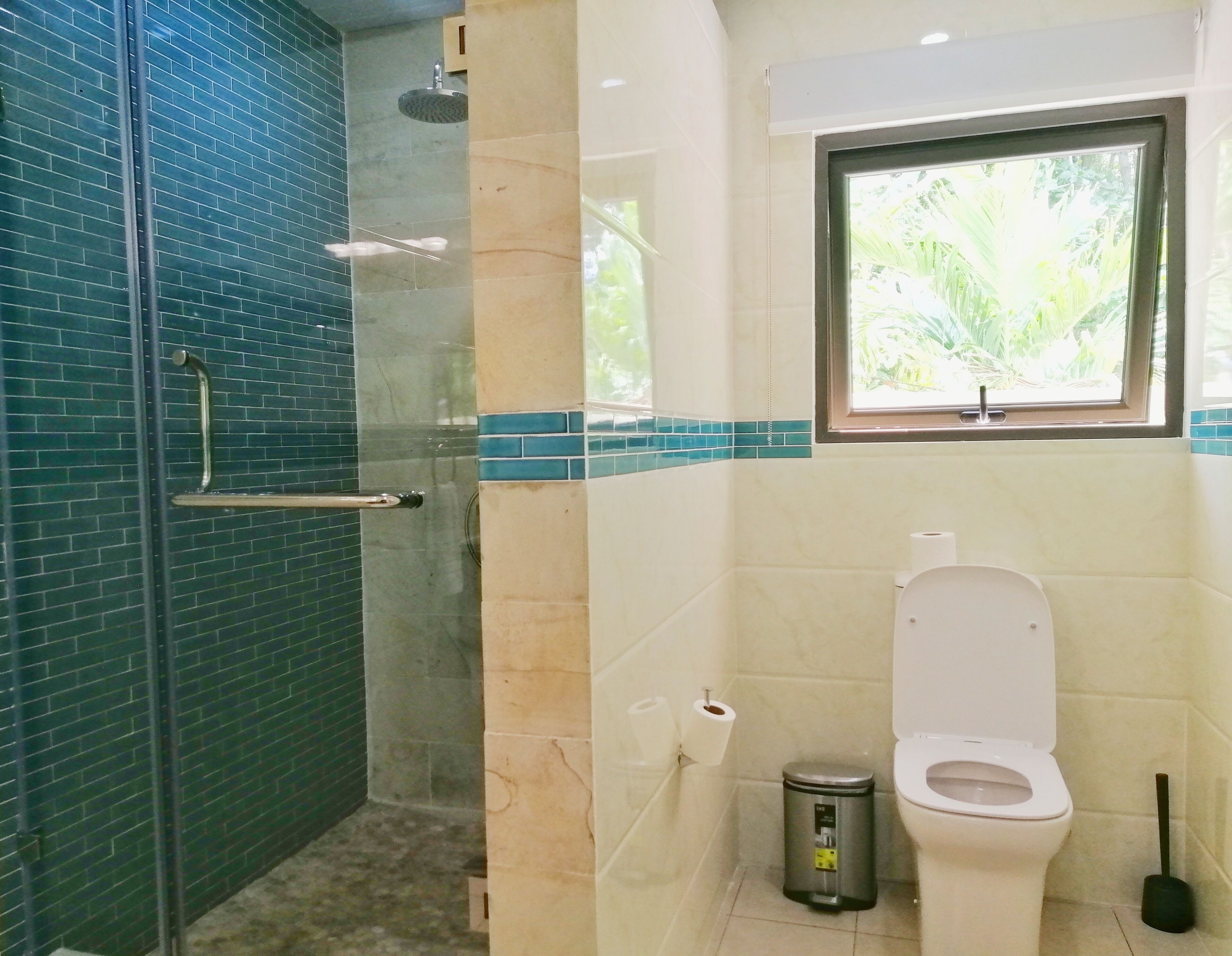 A bathroom with a glass-enclosed shower, a toilet, and a window with a view of greenery.
