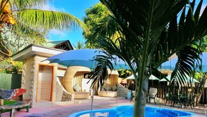 Zona de piscina con sillas de descanso, un pequeño edificio y plantas tropicales.
