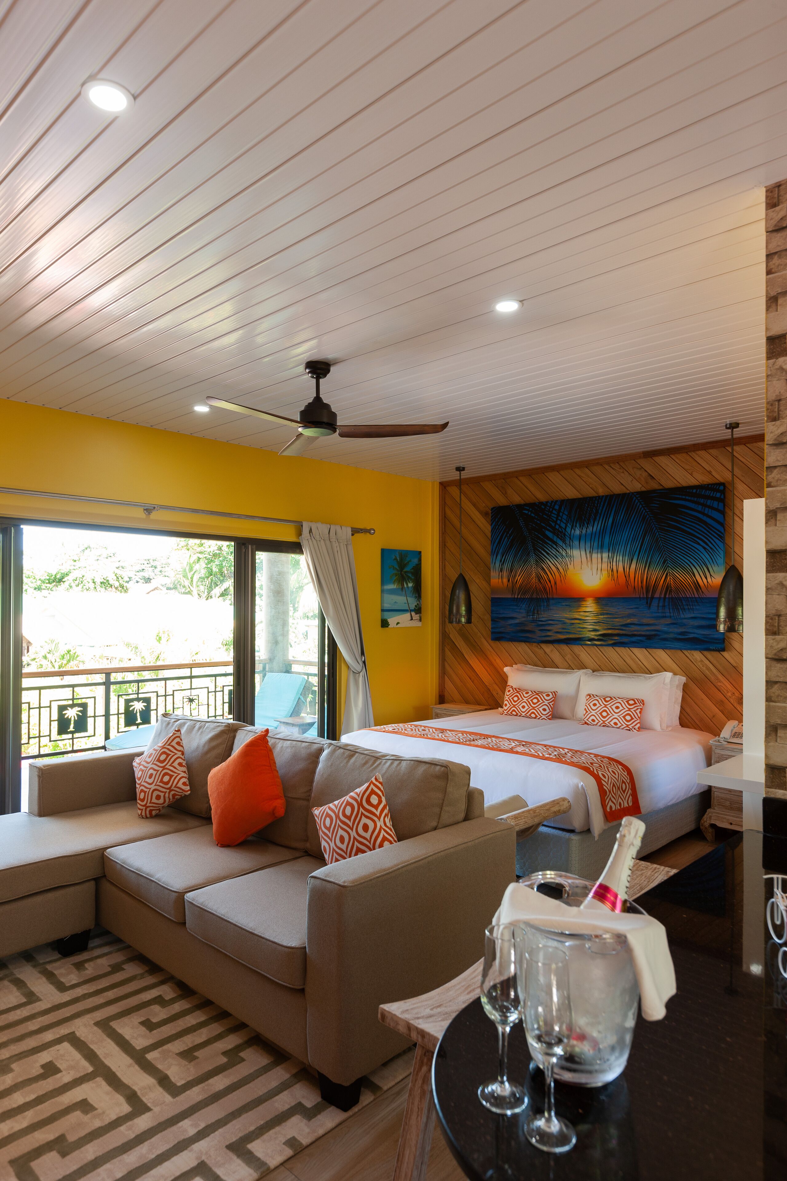A hotel room with a bed, a sofa, a balcony, a painting, and a bottle of champagne.