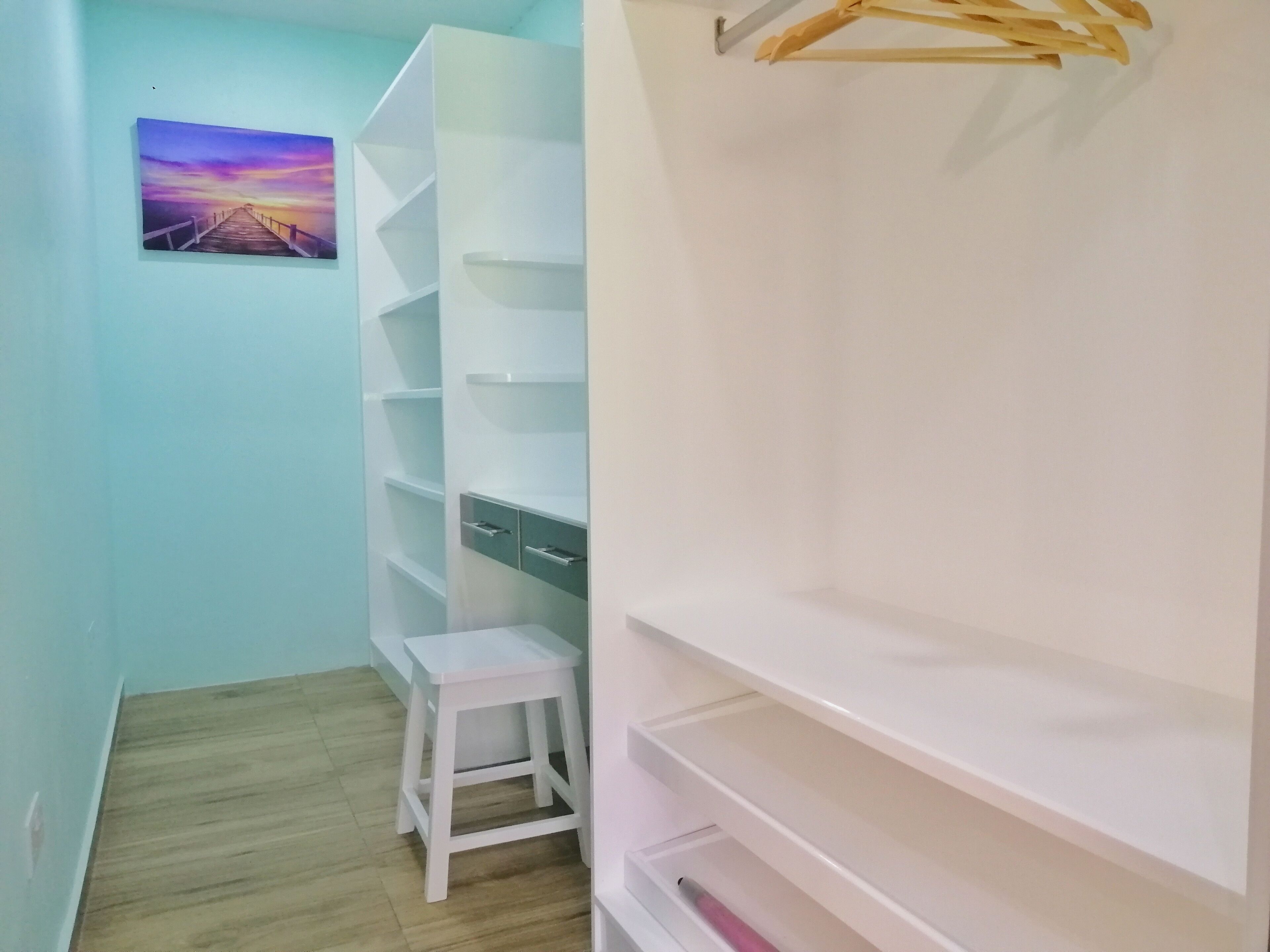A narrow hallway with a wooden floor, a white closet with multiple shelves, a small white stool, and a wooden hanger rack.