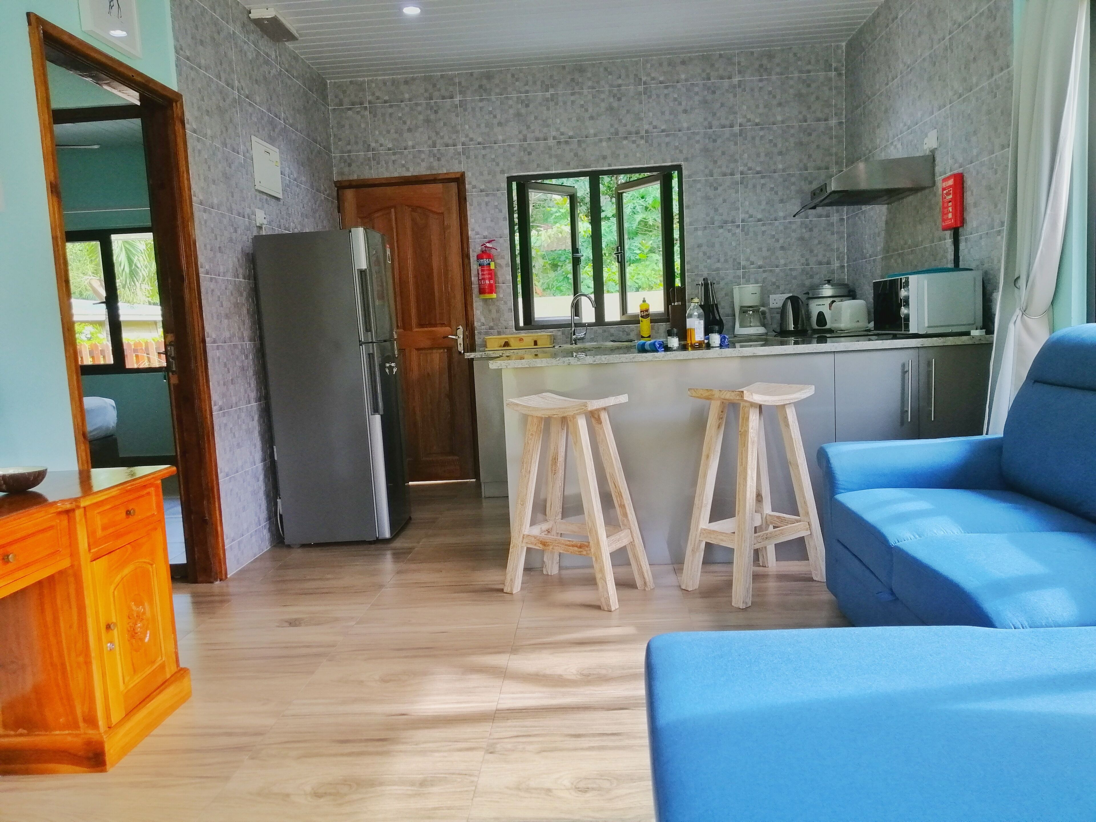 A compact kitchen with a refrigerator, wooden stools, a blue sofa, and a wooden cabinet.