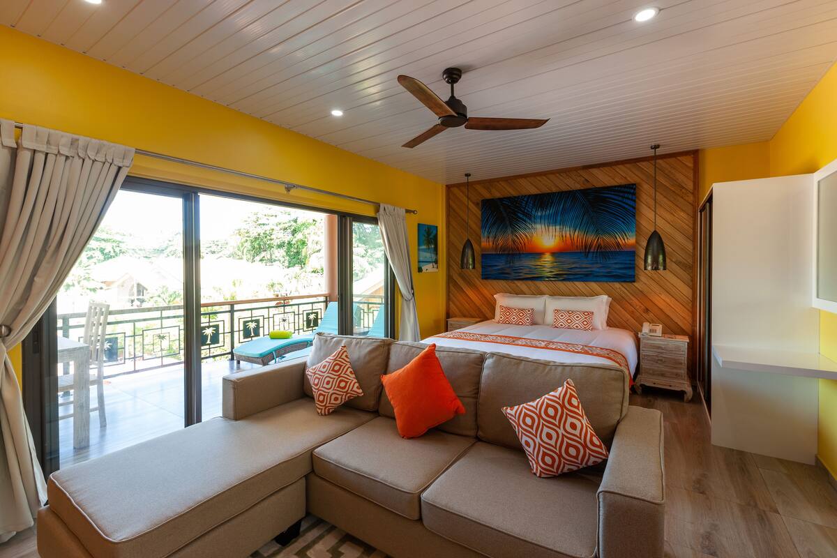 A hotel room with a bed, a sofa with patterned cushions, a ceiling fan, and a painting on the wall.
