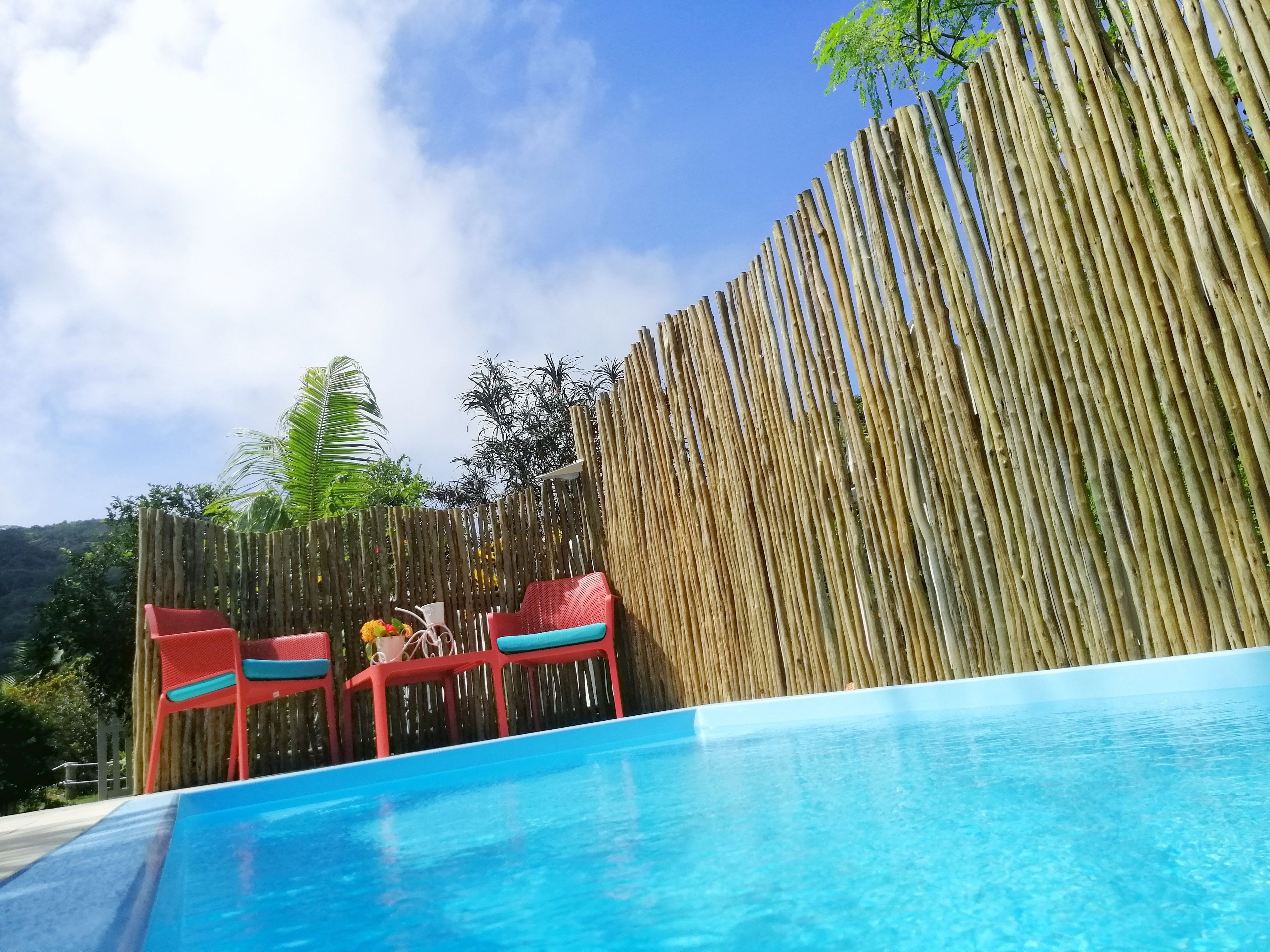 Une piscine entourée d’une clôture en bambou, avec des chaises rouges et un ciel d’un bleu limpide.