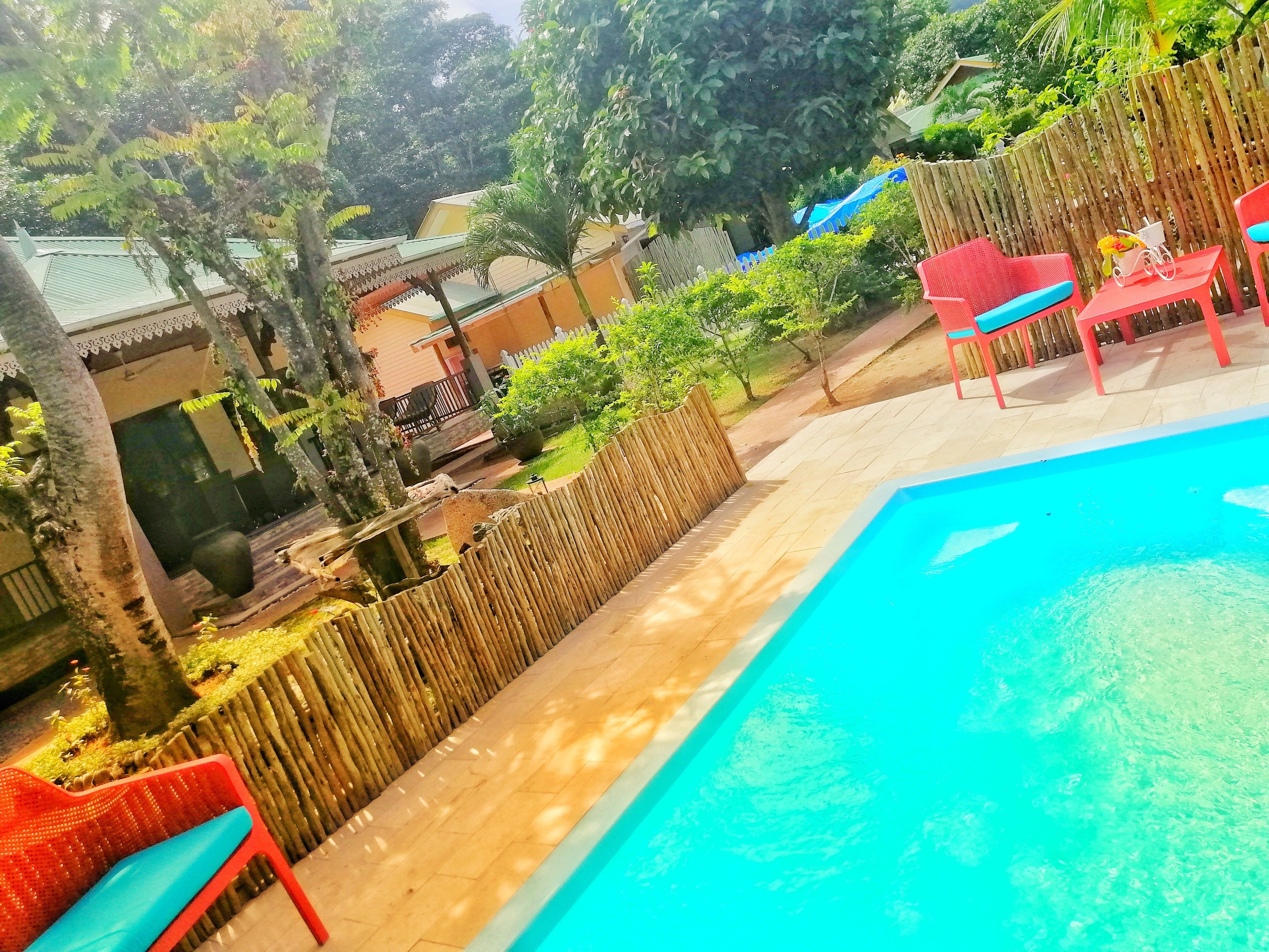 Una piscina con sillas de descanso rojas y azules, una valla de madera y vistas a un edificio con techo verde.
