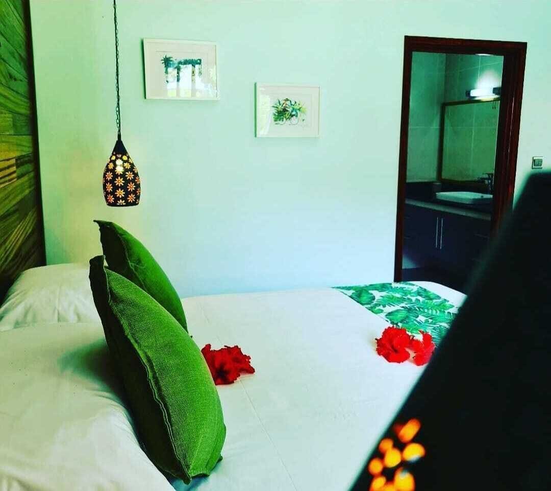 Une chambre d’hôtel avec un lit, des oreillers verts, un luminaire suspendu et deux tableaux encadrés au mur.