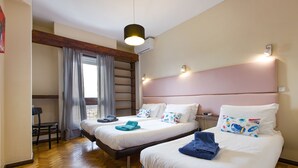 A hotel room with two beds, a wooden floor, a desk, and a window with curtains.