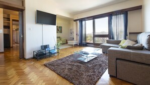 A modern living room with a sofa, a coffee table, and a TV on a stand. There is a large window with curtains, a wooden floor, and a door leading to another room.