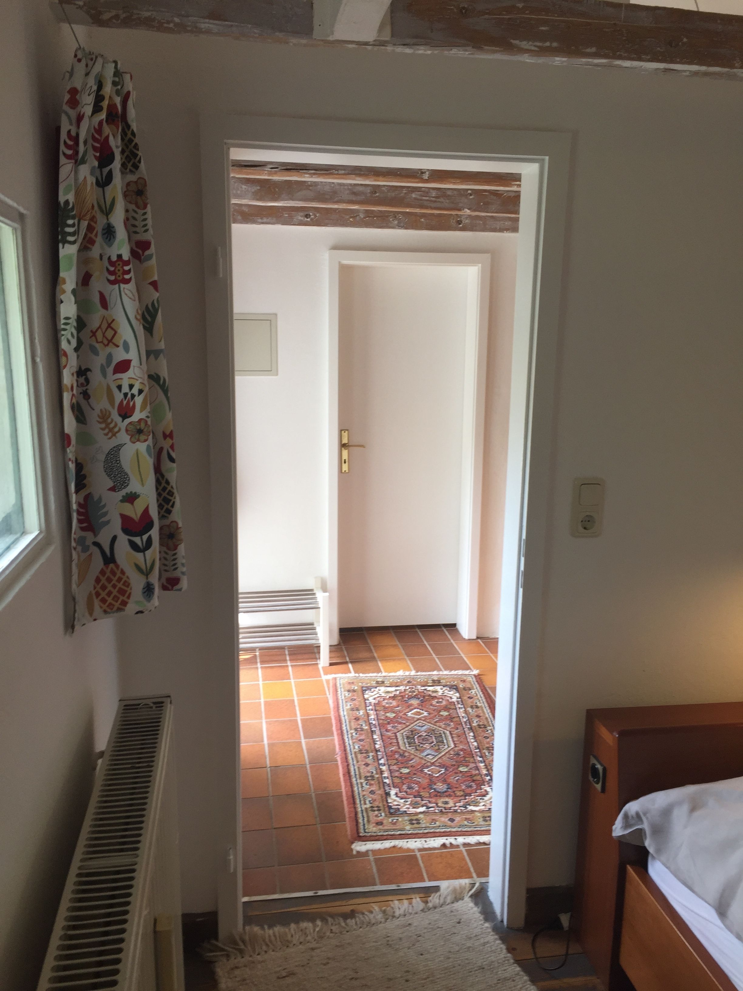 A narrow hallway with a door leading to a room with a bed and a radiator. A patterned curtain hangs on the left, and a rug is on the floor.