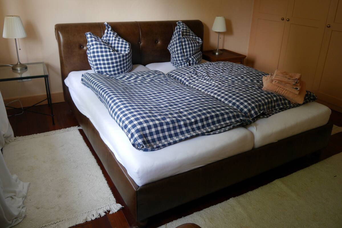 Ein ordentlich gemachtes Bett mit karierter Bettwäsche, ein Nachttisch mit Lampe und ein gefaltetes Handtuch am Fußende.