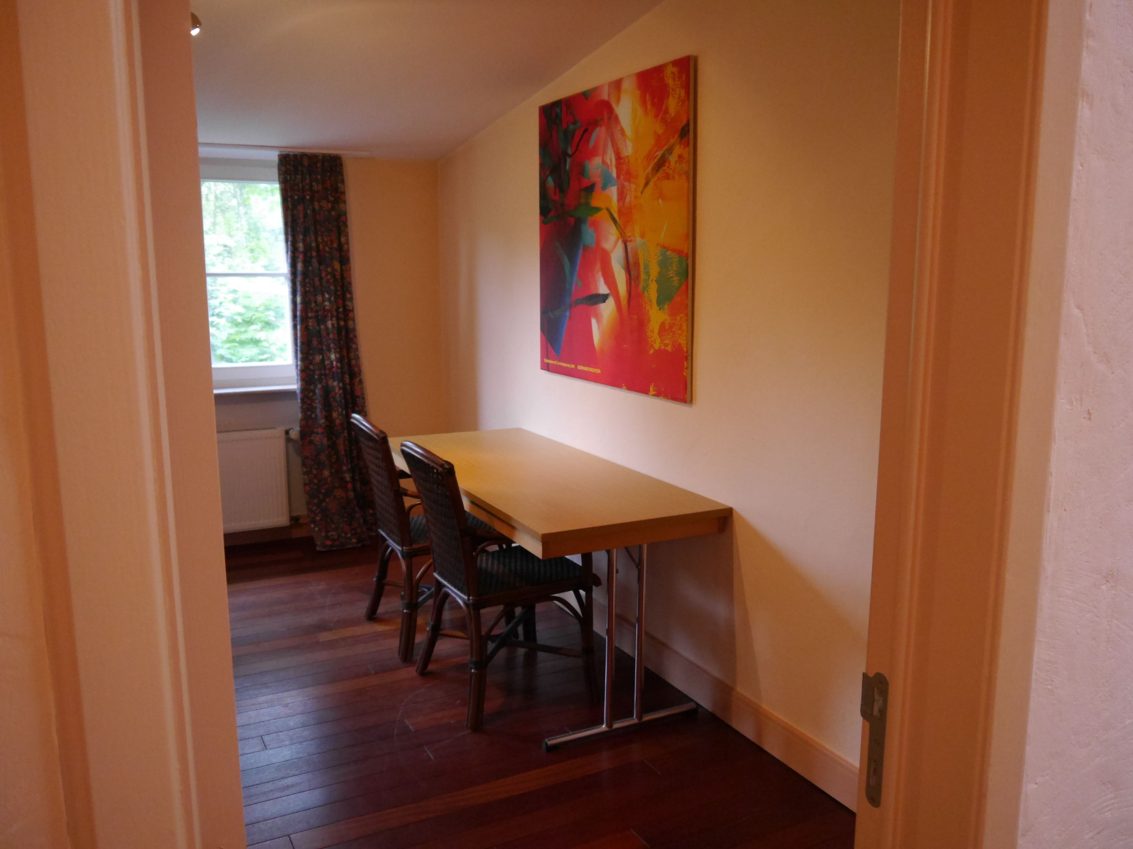 A room with a wooden dining table and chairs, a window with curtains, and a colorful abstract painting on the wall.