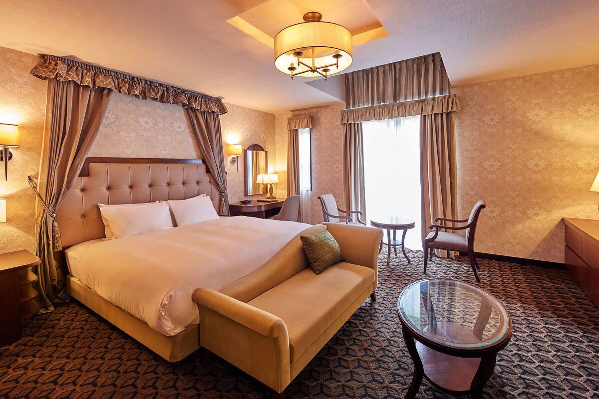 A hotel room with a large bed, a sofa, a small table, and a chair. There is a window with curtains and a lamp on the wall.