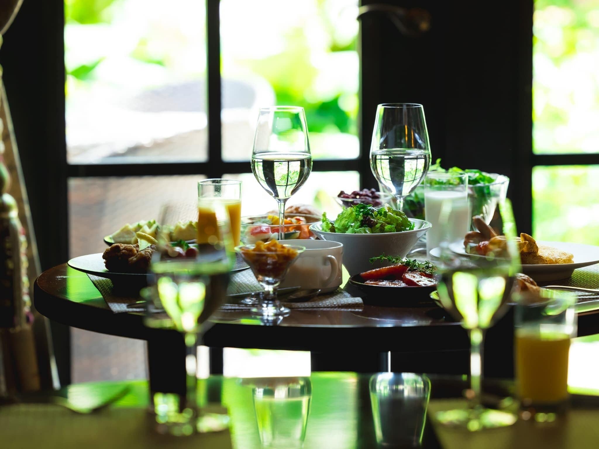 Mesa de jantar posta com diversos pratos e bebidas, próxima a uma janela com vista para o verde.