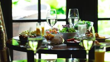 Mesa de jantar posta com diversos pratos e bebidas, próxima a uma janela com vista para o verde.