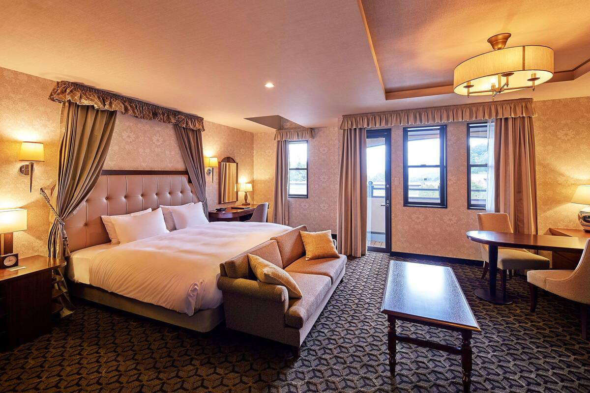 A hotel room with a large bed, a sofa, a dining table, and a chandelier.