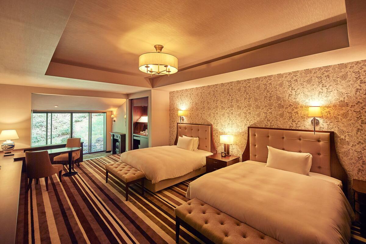 A hotel room with two beds, a seating area, and a view of the outdoors.