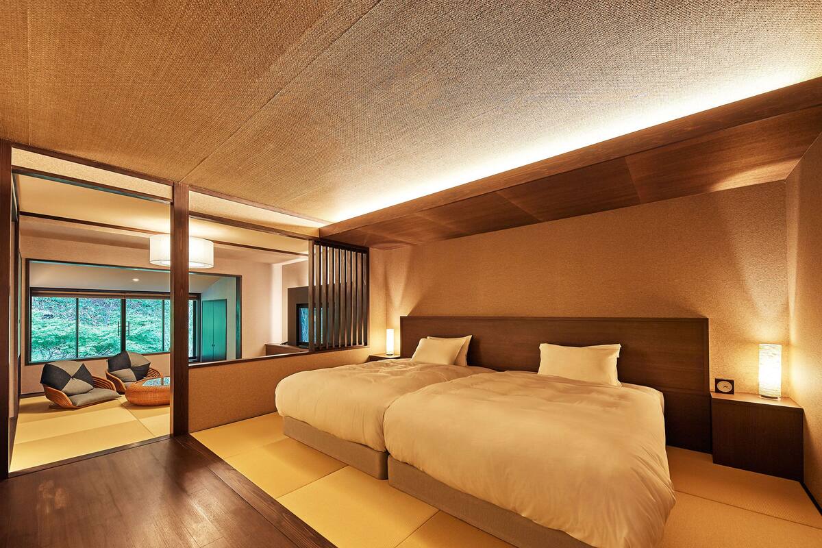 A modern hotel room with a large bed, a nightstand, and a view of a forest through the window.