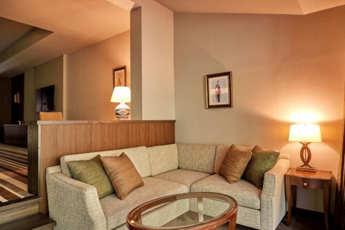 A hotel room with a sofa, a glass coffee table, a lamp, and a framed picture on the wall.