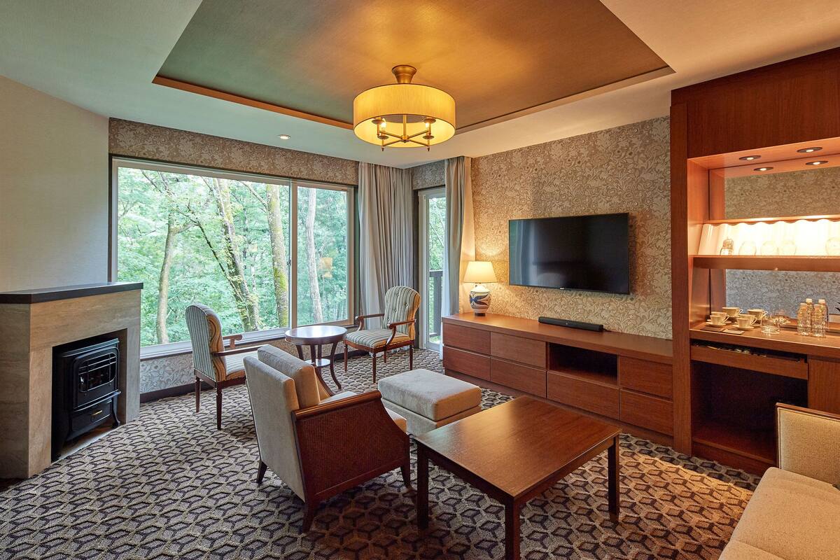 A modern hotel room with a fireplace, a television, a dining area, and a large window offering a forest view.