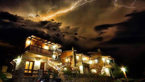 A two-story stone building with a balcony, a motorcycle parked in front, and lightning in the sky. - Efharis Filoxenon (Sikyona)
