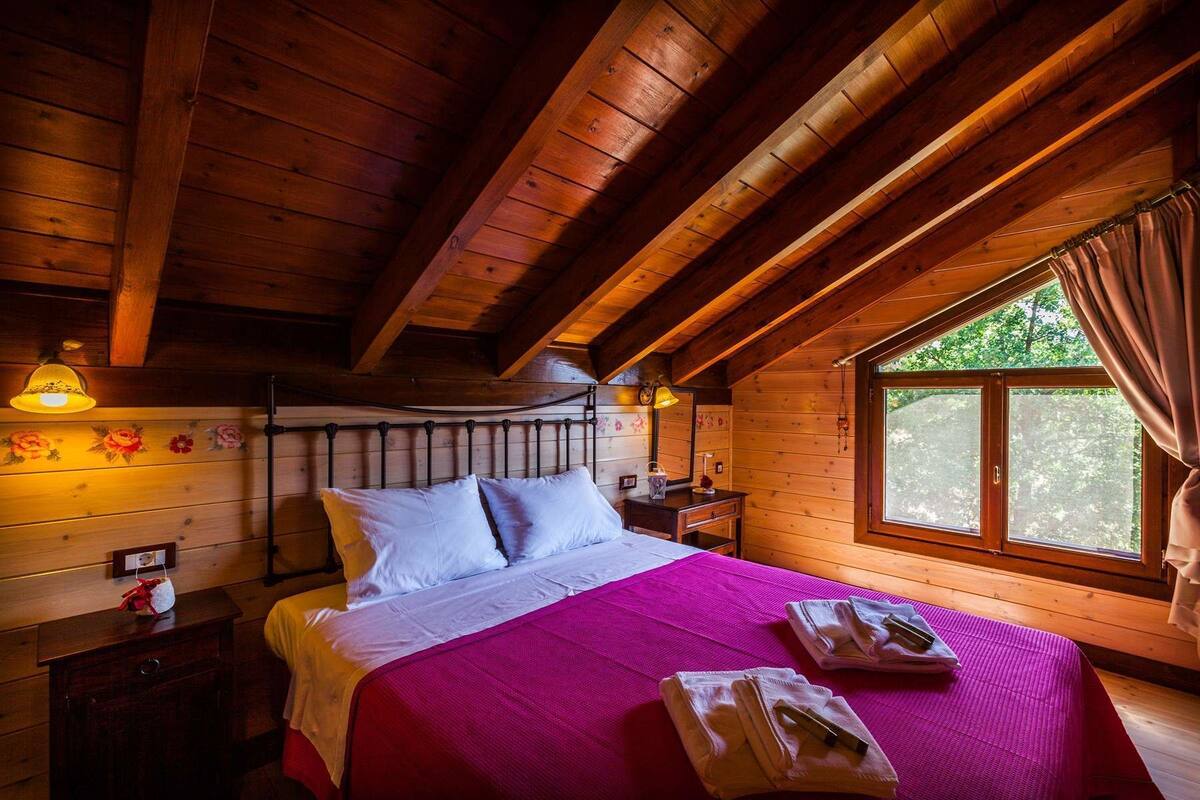 A wooden cabin room with a bed, a nightstand, a dresser, and a window with curtains.