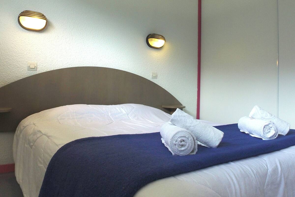 A neatly made bed with white linens and a blue blanket, two rolled towels, and two wall-mounted lights.