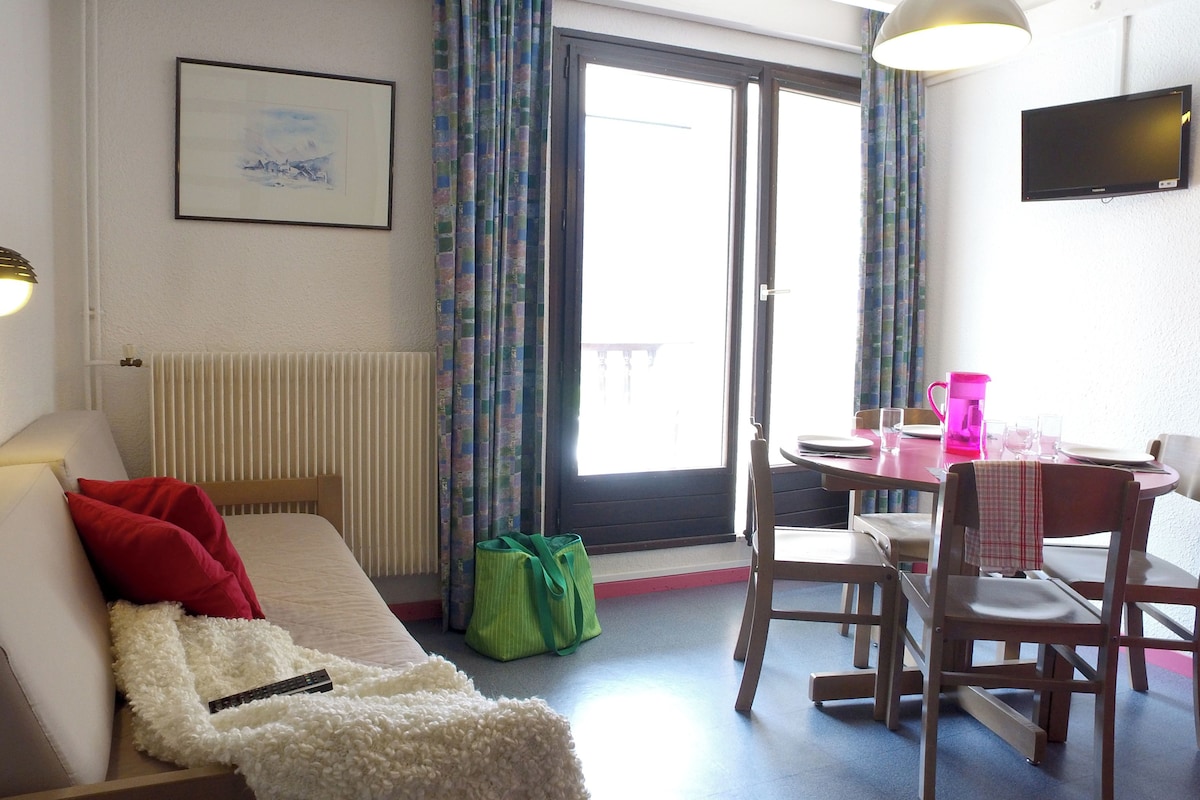 A hotel room with a bed, a small table with chairs, a radiator, a window with curtains, and a flat-screen TV mounted on the wall.