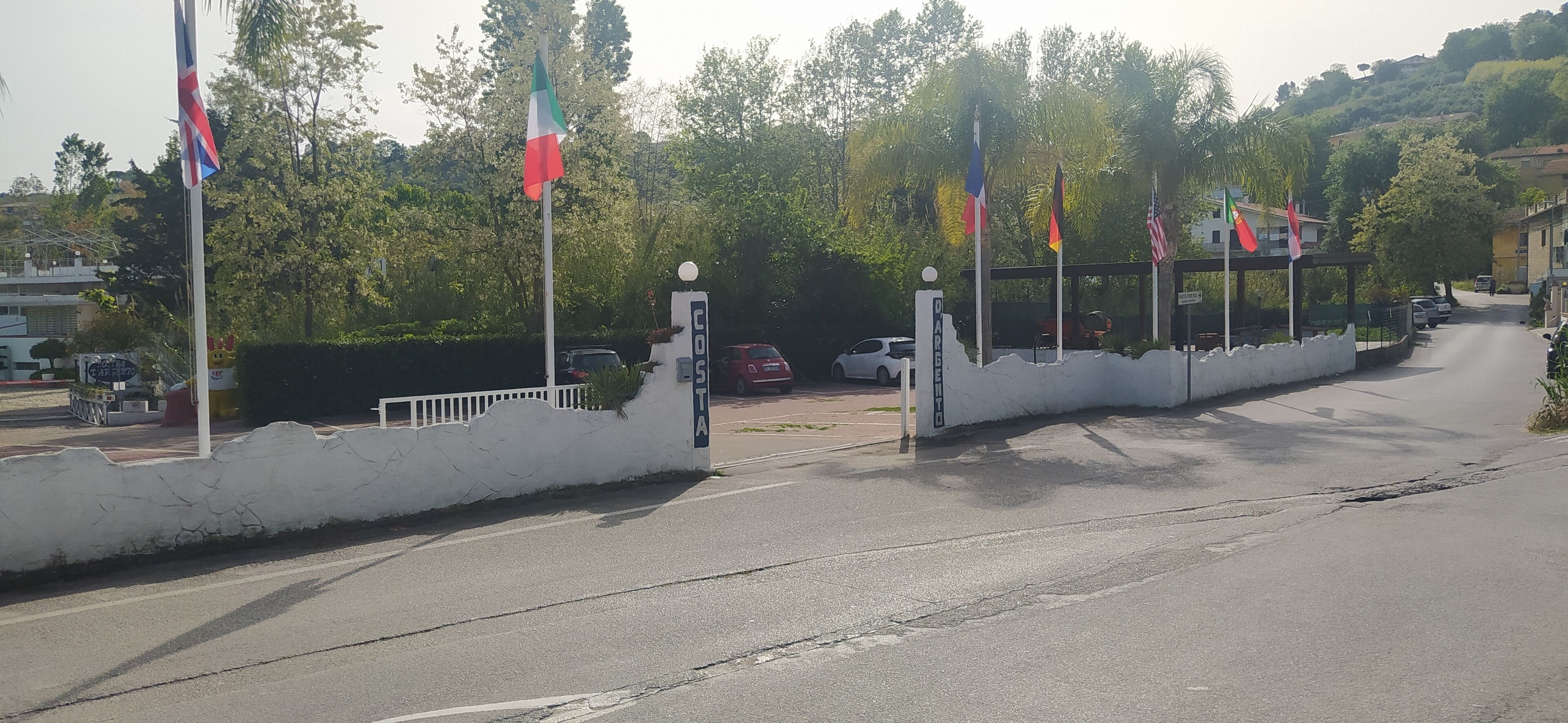 A street with multiple flags, a white wall, and parked cars.