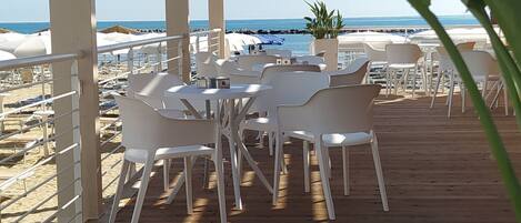 Terraza de madera con sillas y mesas blancas, con vista a una playa.