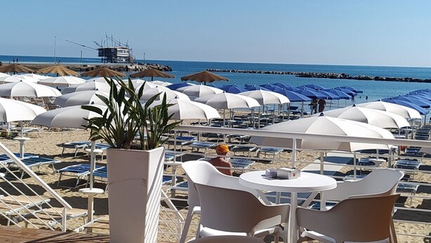 Een terras aan het strand met witte plastic stoelen en tafels, een plant in pot, en uitzicht op het strand en de oceaan.