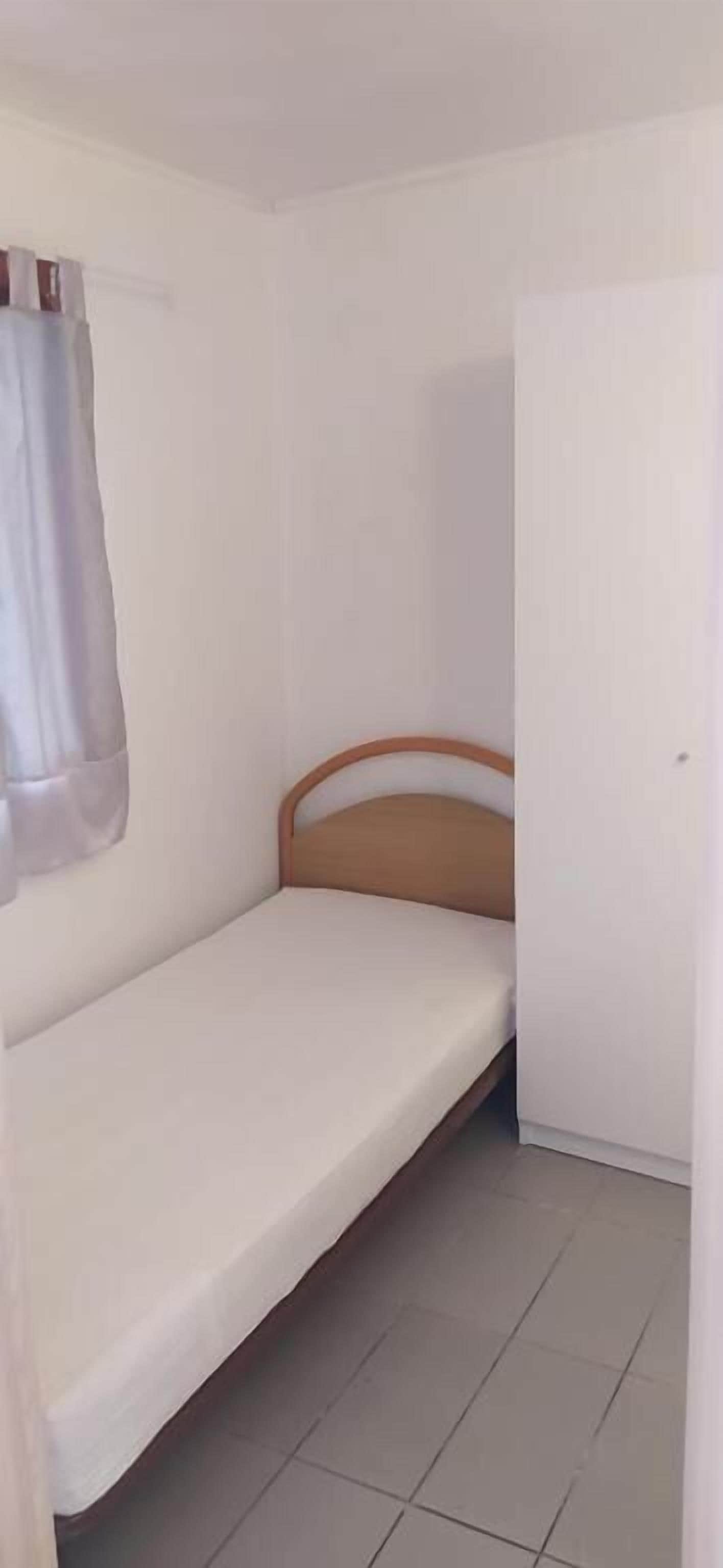 Une petite chambre avec un lit simple, une tête de lit en bois, une porte blanche et un rideau.