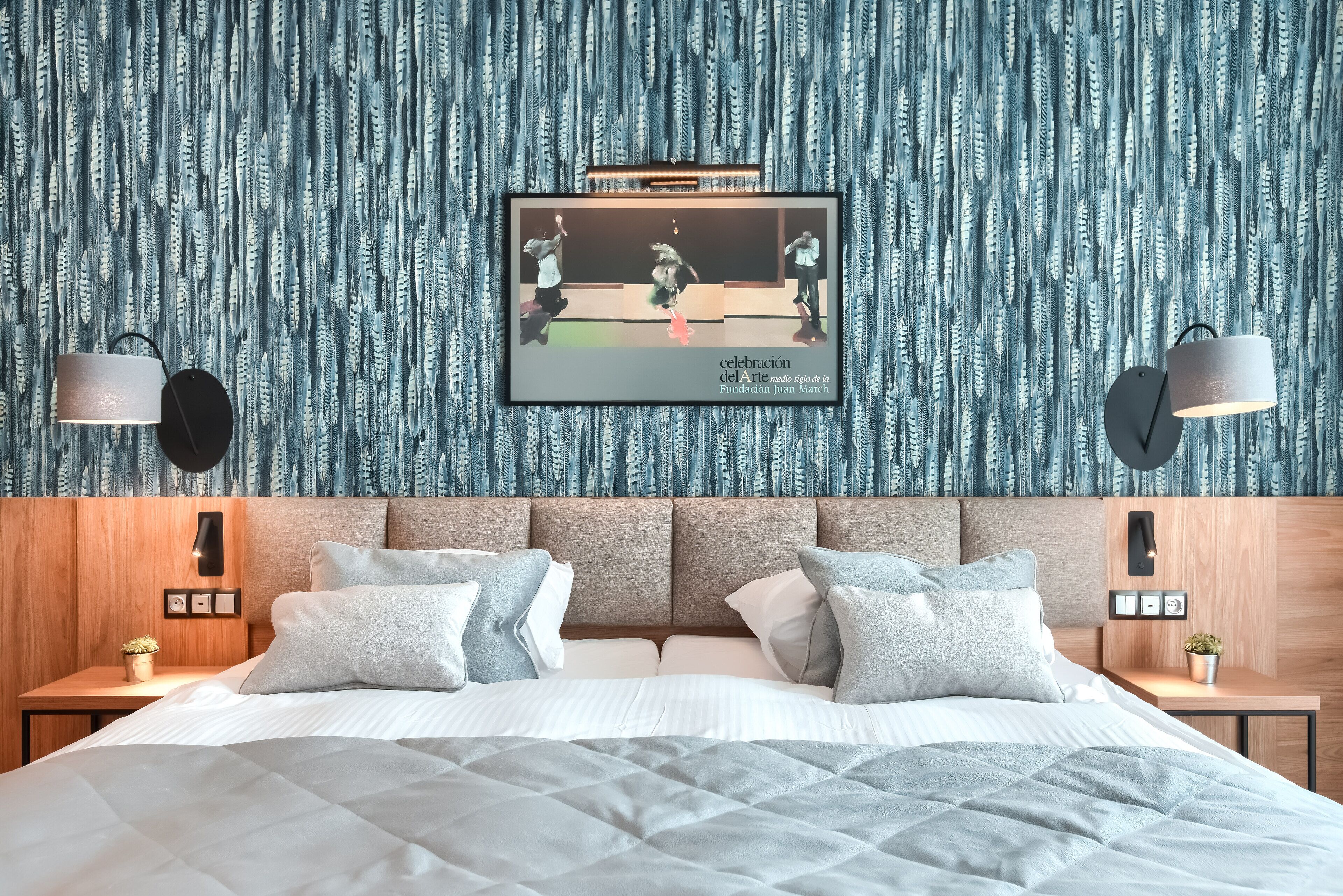 Un dormitorio con cama, almohadas, cabecero, mesitas de noche, lámparas de pared y una obra de arte enmarcada que representa un combate de boxeo.
