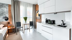 Een moderne hotelkamer met een bed, een klein bureau met lamp, een stoel, een eettafel met twee stoelen, een keuken met een spoelbak en kastjes, en een groot raam met gordijnen.