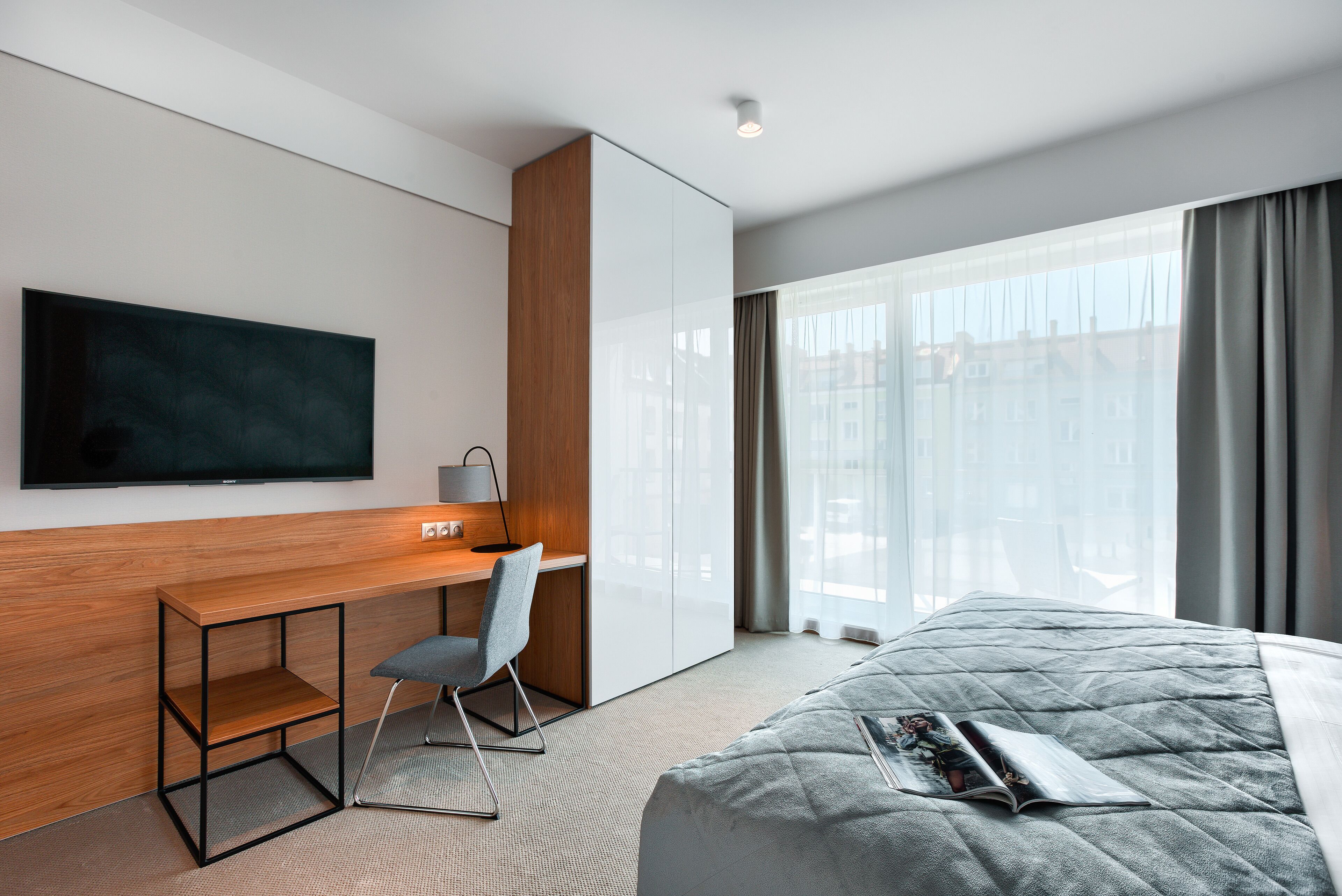 Een moderne hotelkamer met een groot bed, een bureau met stoel, een flatscreen televisie aan de wand en een groot raam met gordijnen.