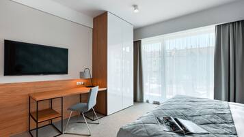 A modern hotel room with a large bed, a desk with a chair, a flat-screen TV mounted on the wall, and a large window with curtains.