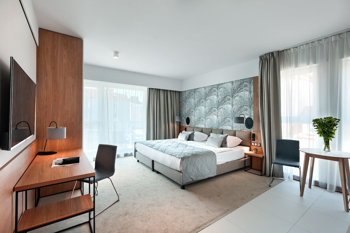 A modern hotel room with a large bed, a desk, a chair, a TV, and a plant.