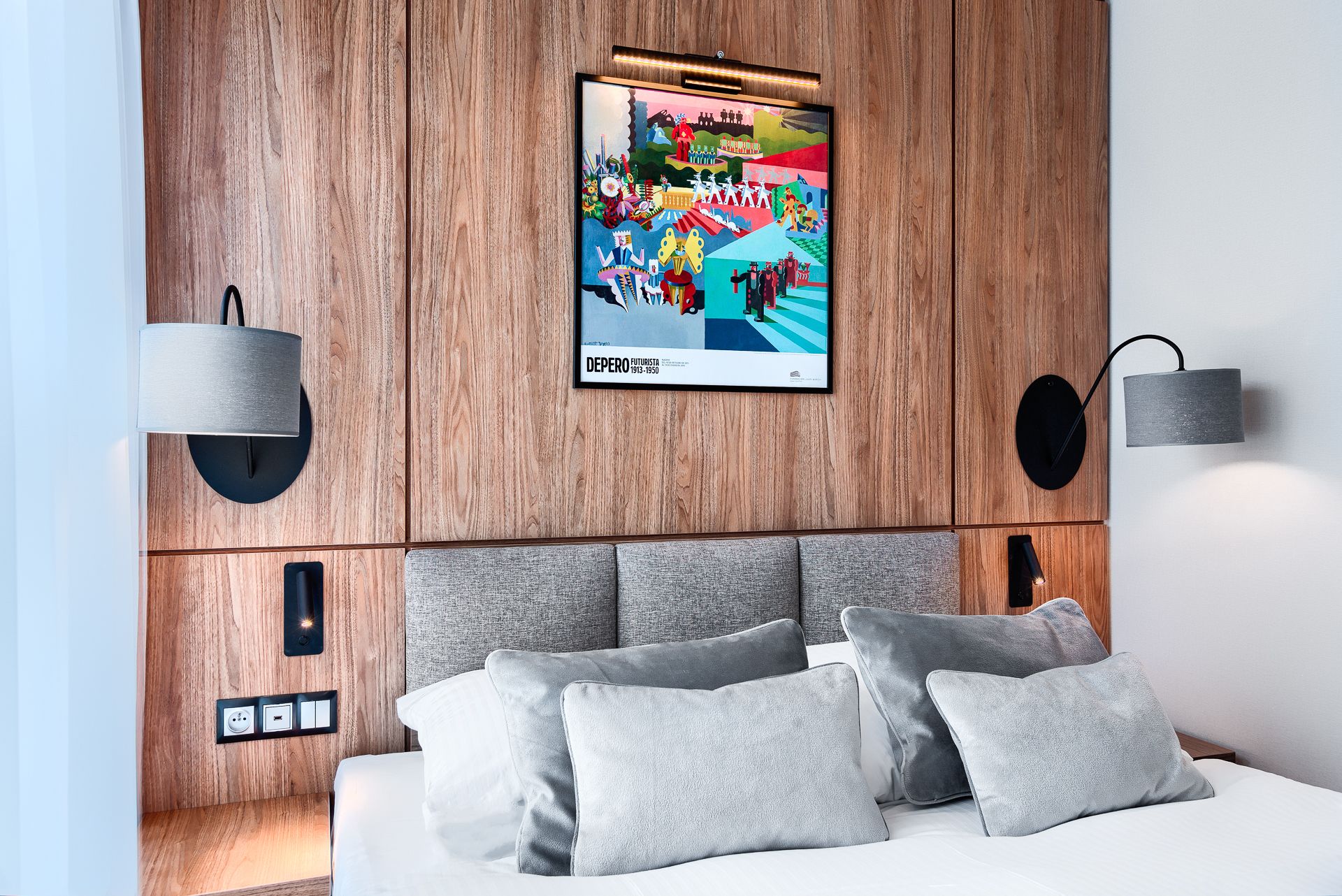 Une chambre d’hôtel moderne avec une tête de lit en bois, une grande affiche représentant « DBEPERO FUTURISTA 1913-1950 » et un lit avec des oreillers gris.