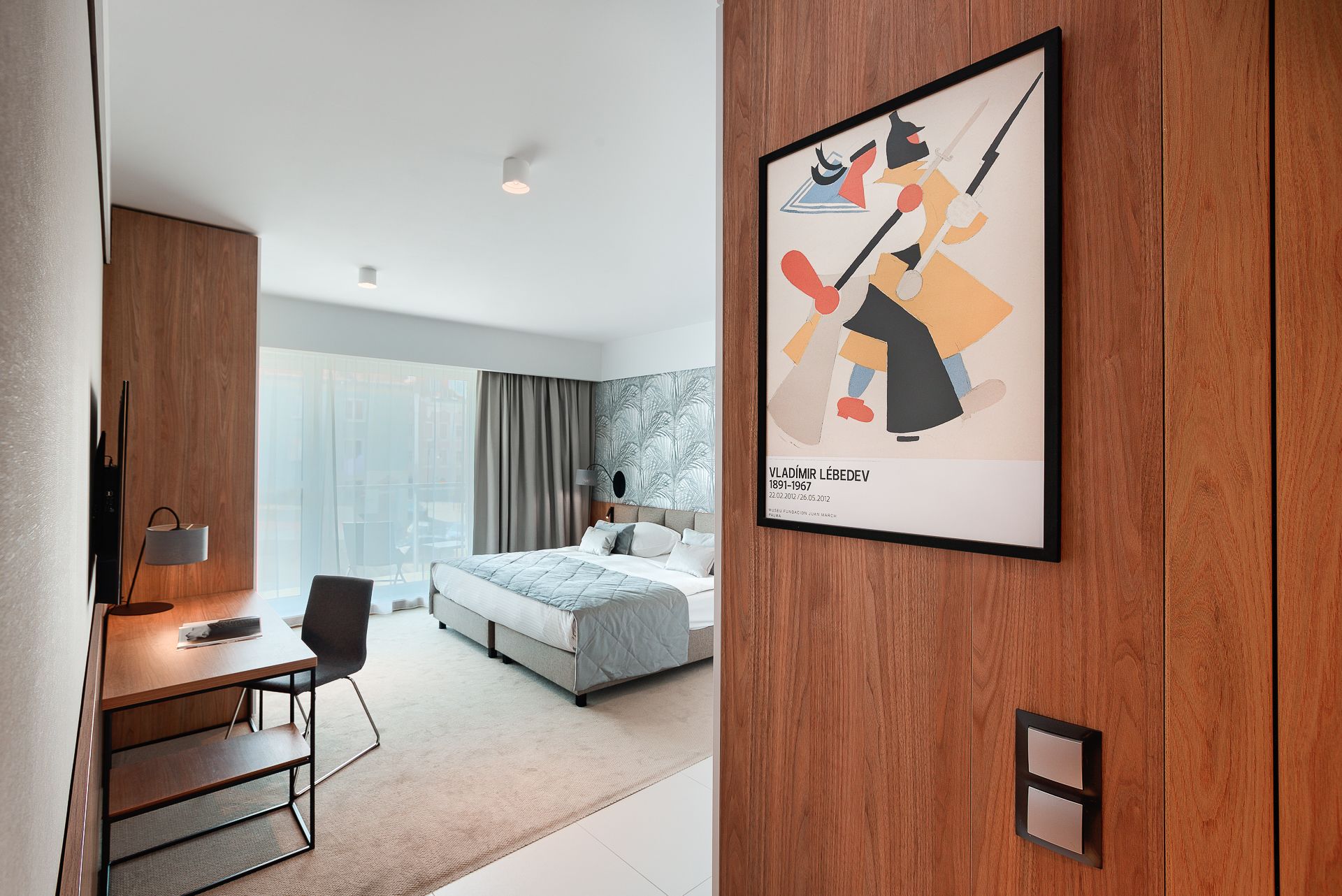Una habitación de hotel moderna con cama, escritorio y silla, un ventanal grande y una obra de arte enmarcada en la pared.