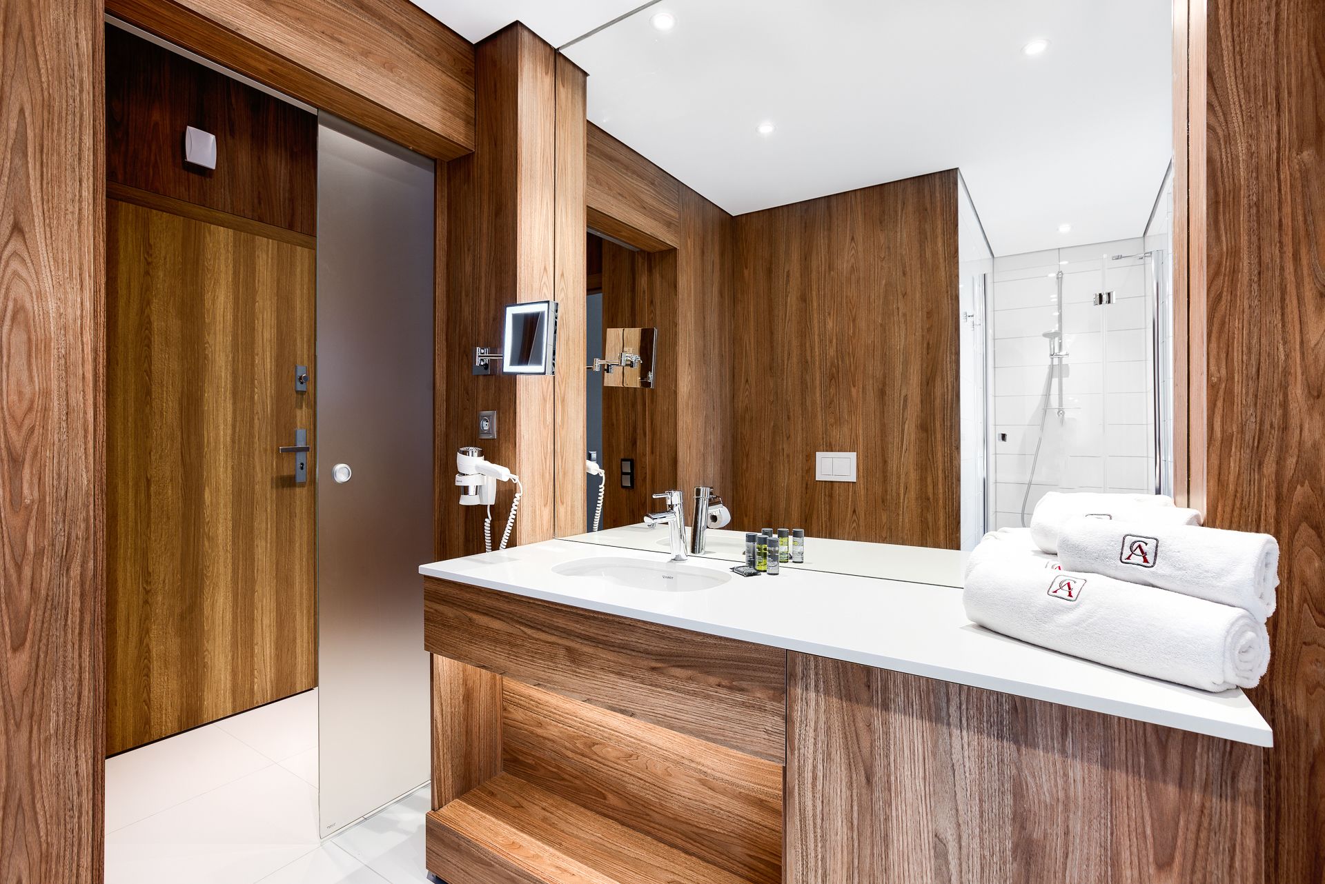 A modern bathroom with wooden paneling, a large mirror, a shower, and a vanity with a towel rack.