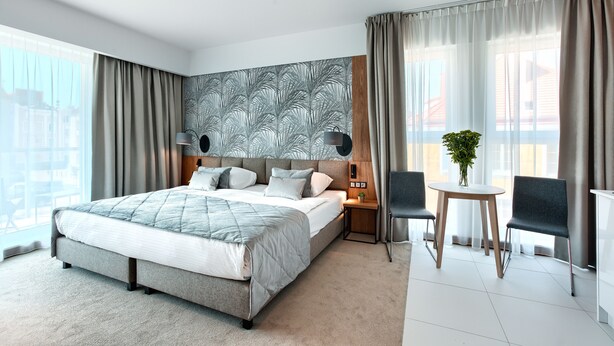 Een moderne hotelkamer met een groot bed, een nachtkastje met lamp, een kleintje eettafeltje met stoelen, en uitzicht op de stad door grote ramen.
