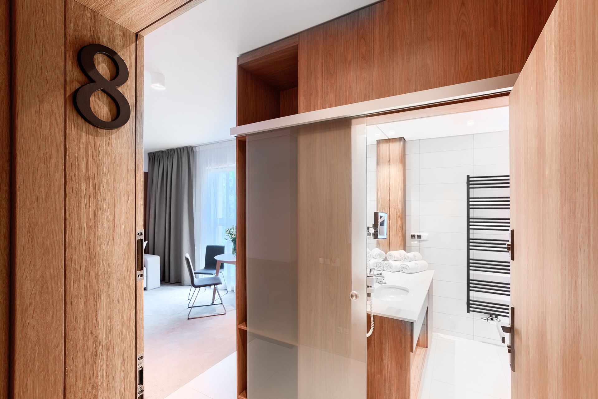 A modern hotel room with a wooden wardrobe, a bathroom with a towel rack, and a dining area with a table and chairs.