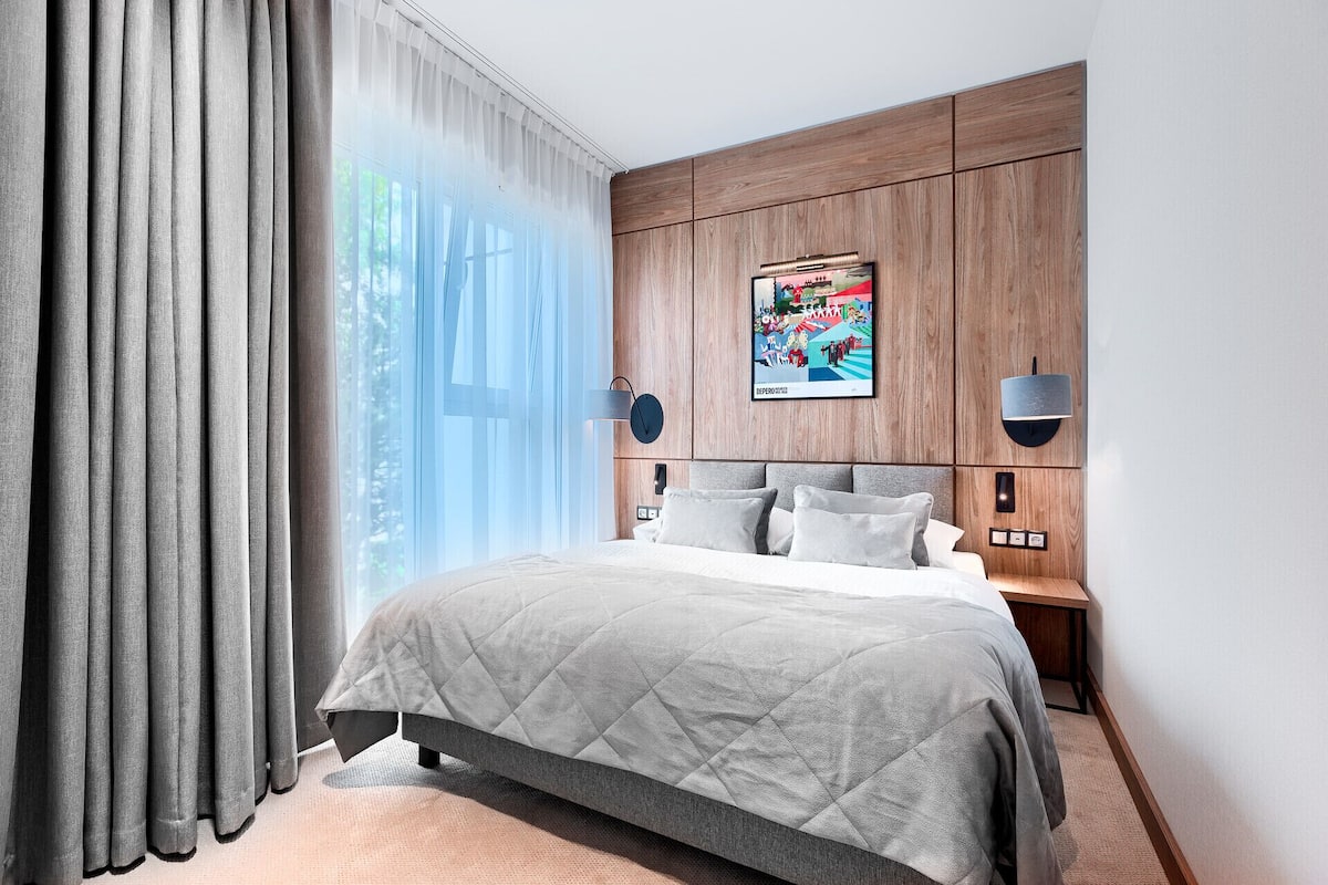 A modern hotel room with a large bed, a wooden headboard, a wall-mounted TV, and a large window with curtains.