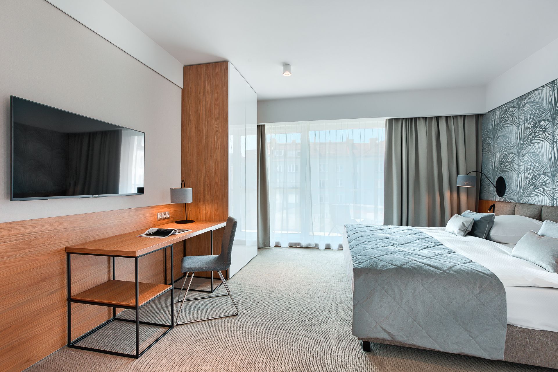 A modern hotel room with a bed, a desk, a chair, a TV, and a large window with curtains.