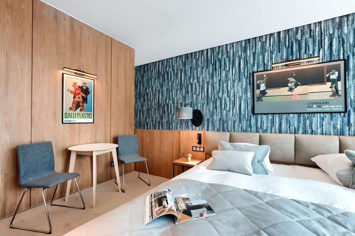 A hotel room with a bed, a small table, two chairs, a nightstand, and a poster on the wall.