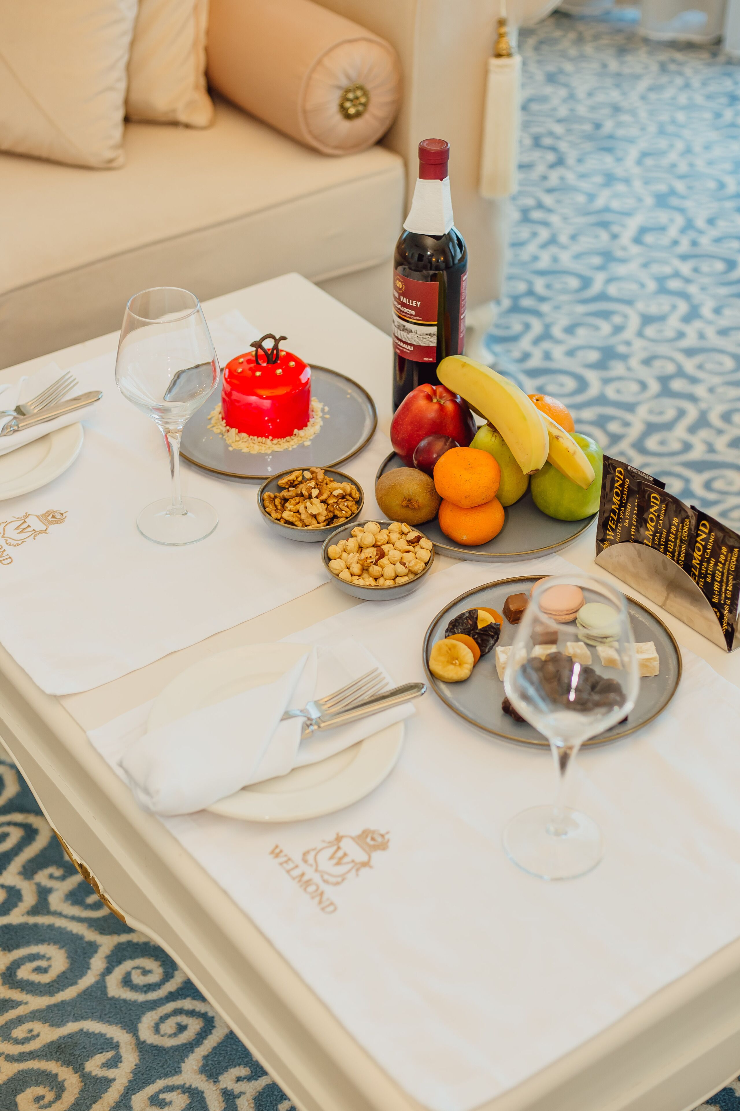 A dining table set with plates, cutlery, wine glasses, a bottle of wine, a red cake, fruits, nuts, and macarons.