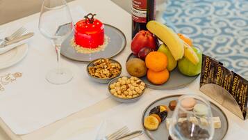 A dining table set with plates, cutlery, wine glasses, a bottle of wine, a red cake, fruits, nuts, and macarons.
