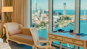 A hotel room with a large window offering a city view, a sofa, a desk with a phone and a book, and a lamp. - JRW Welmond Hotel & Casino Batumi (Batumi)