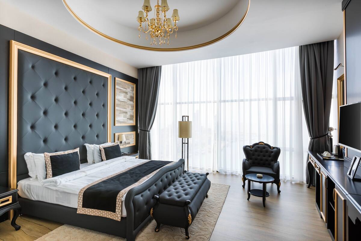 Ein luxuriöses Schlafzimmer mit einem großen Bett, einer Bank, einem Sessel, einem Fernseher und einem großen Fenster mit Vorhängen.