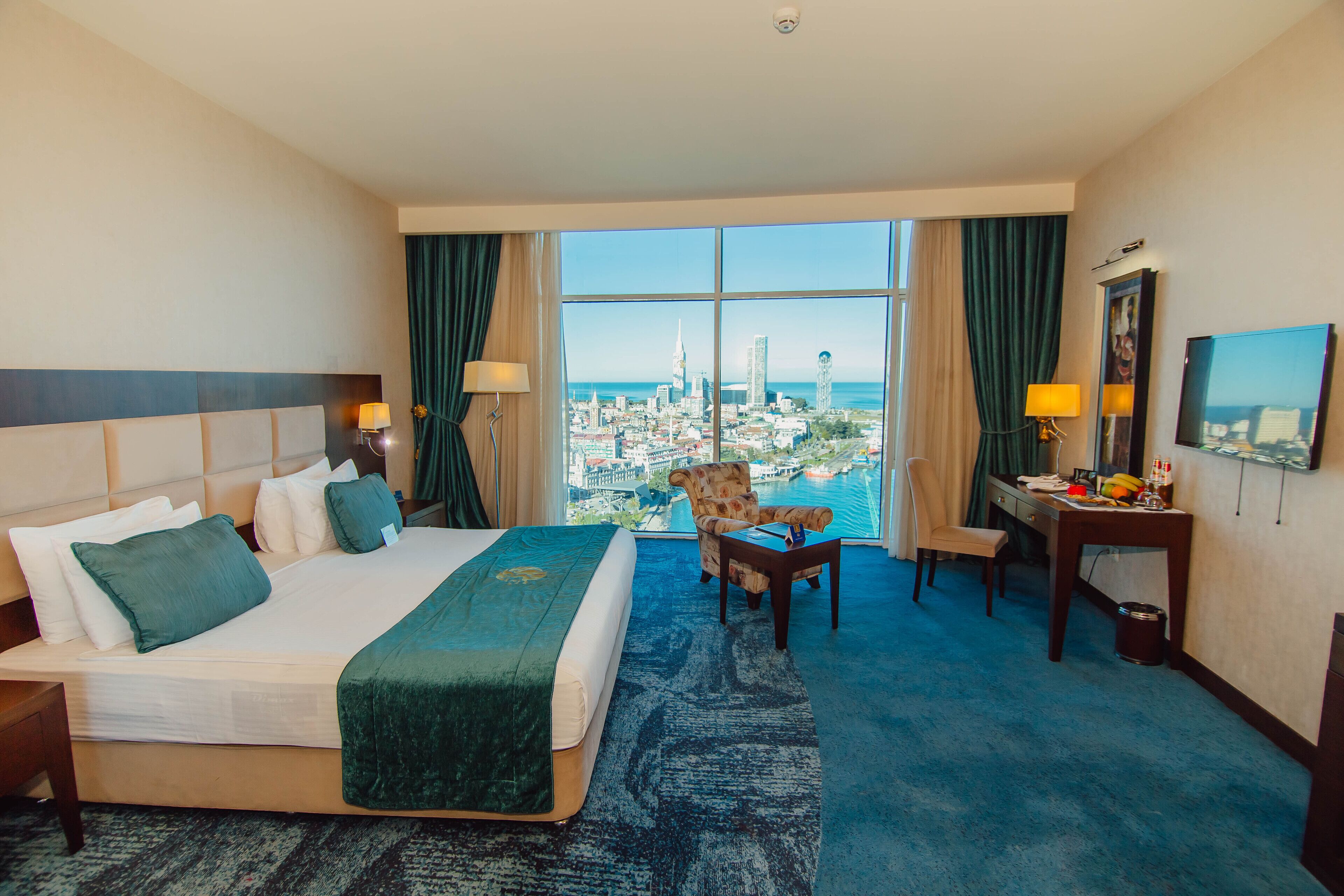 A hotel room with a large bed, a desk, a chair, and a view of a cityscape through large windows.