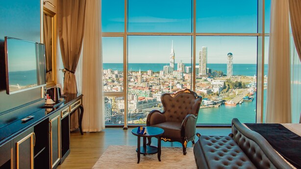 A hotel room with a large window offering a city view, a sofa, a small table, and a TV on a cabinet. - JRW Welmond Hotel & Casino Batumi (Batumi)